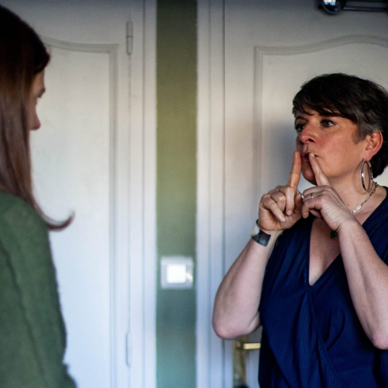 Sandrine est debout en train de faire une séance de sophrologie, elle porte ses mains vers sa bouche pour montrer à la consultante comment il faut expirer. La consultante est face à elle. Il y a une porte blanche derrière elles