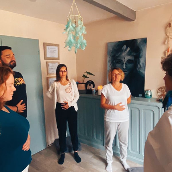 Groupe de 5 personnes en train de faire un exercice de sophrologie respiration abdominale. Les personnes sont debout avec une main sur le ventre. On voit au plafond un attrape reve vert et au fond un tableau d'une femme chamane et des objets posés sur un meuble. 3 cadres accrochés au mur